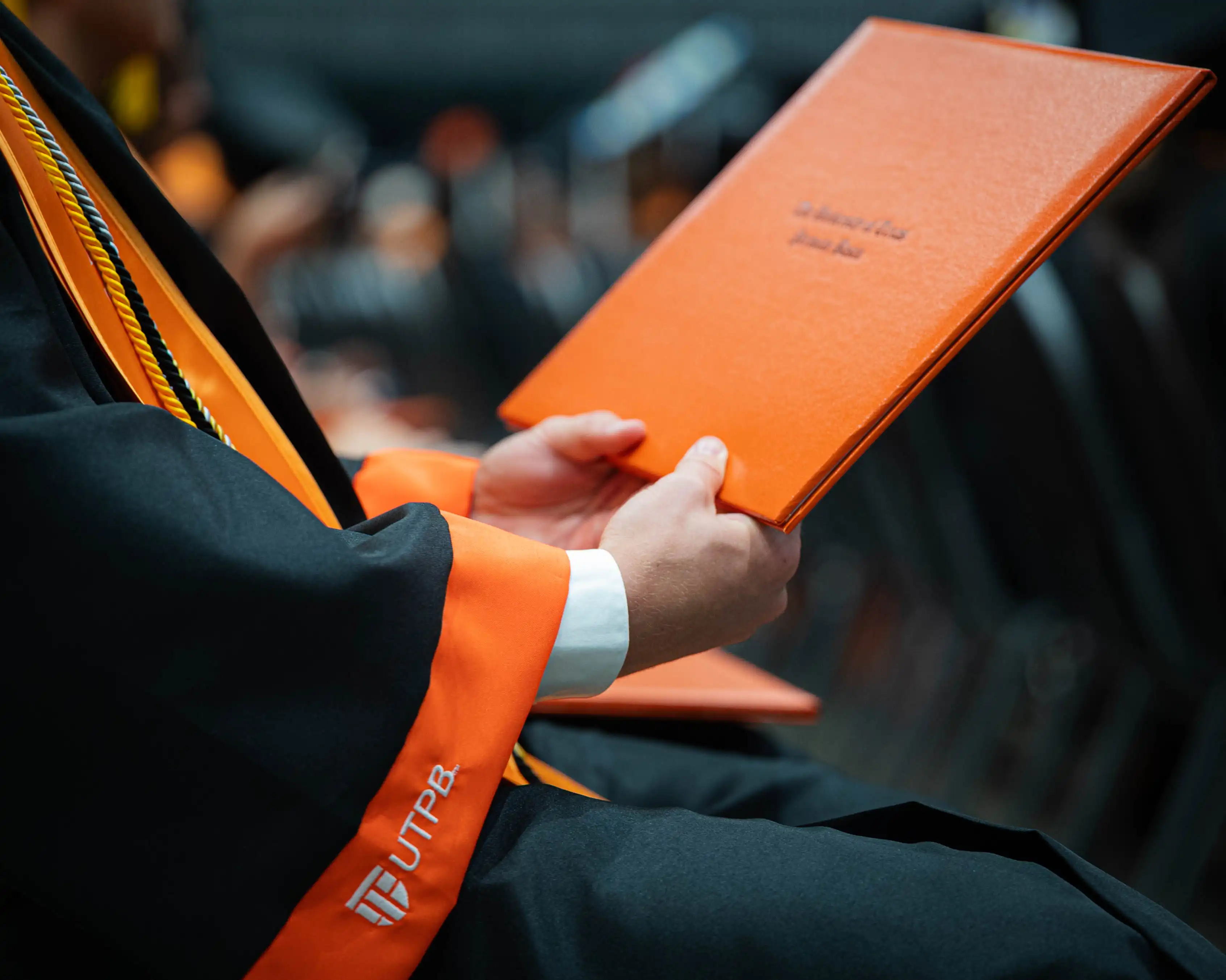 A graduate at graduation admiring their diploma holder in their hands with their graduation regalia on, both inscripted with UTPB.