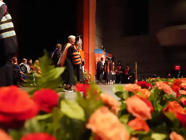A stage decorated for graduation with orange flowers and flags, the president of UTPB congratulating a student on receiving their degree and graduating as the student walks the stage.
