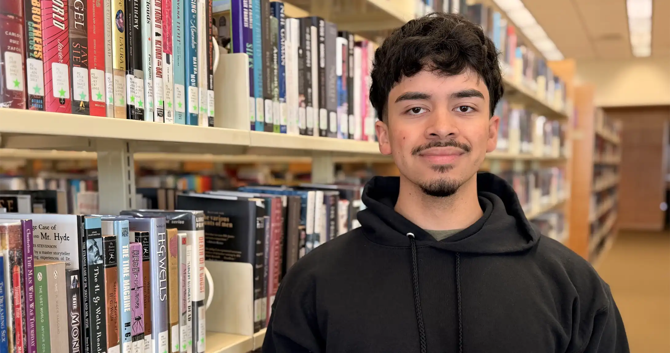 Cesar Vasquez standing near books in the UTPB library.