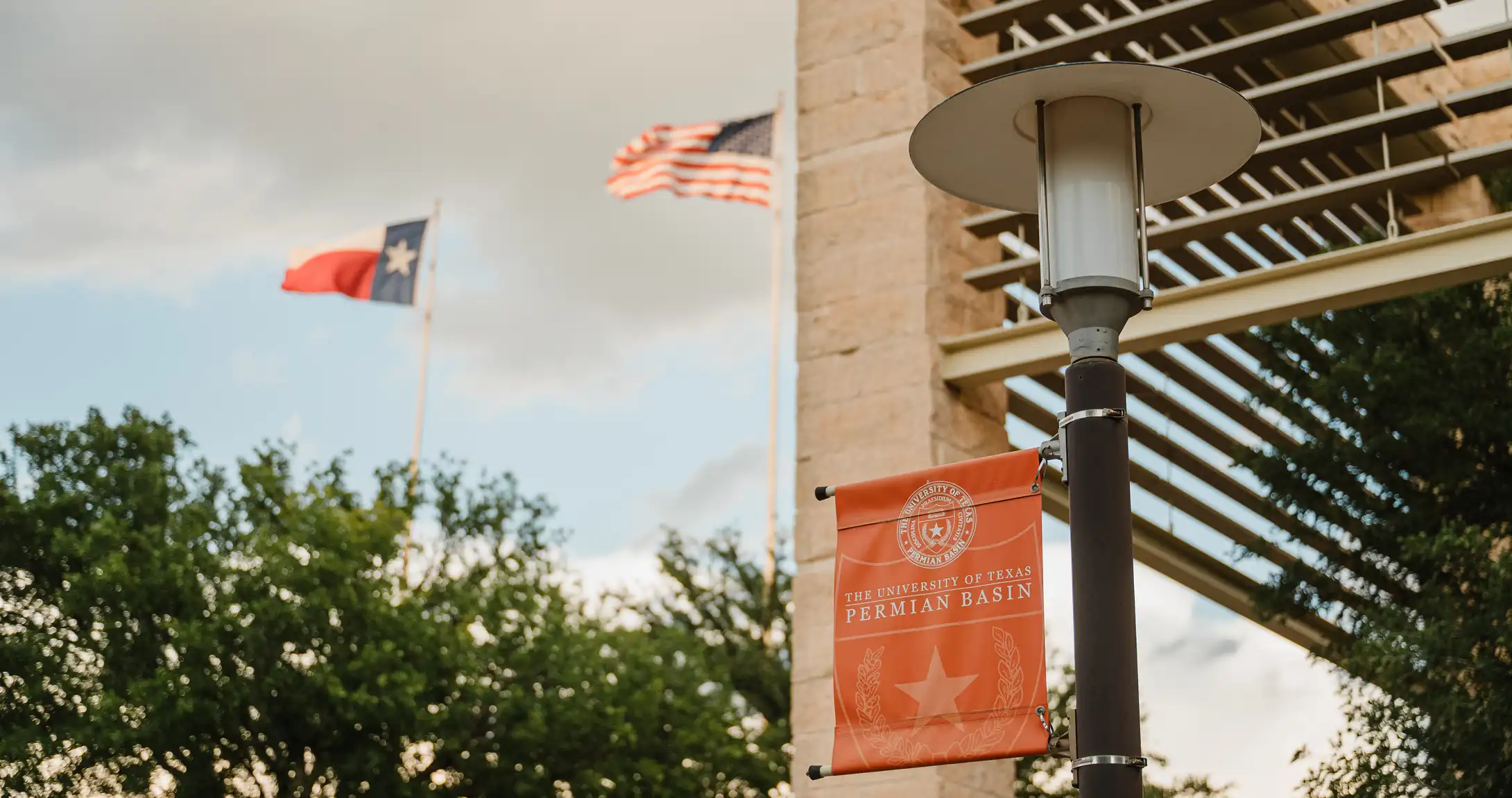 A UTPB flag at the top of a light post with the American and Texas flags in the background. 
