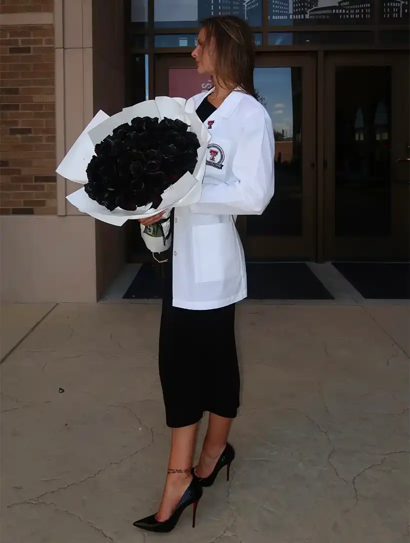Heather wearing a white coat and holding a bouquet of black roses.